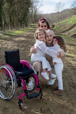 two women and two children crouched on a dirt path.  Pink and black wheelchair in the foreground