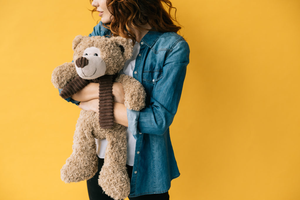 yellow background.  Woman in blue denim jacket, white shirt and black jeans hugging a brown teddy bear with a dark brown scarf