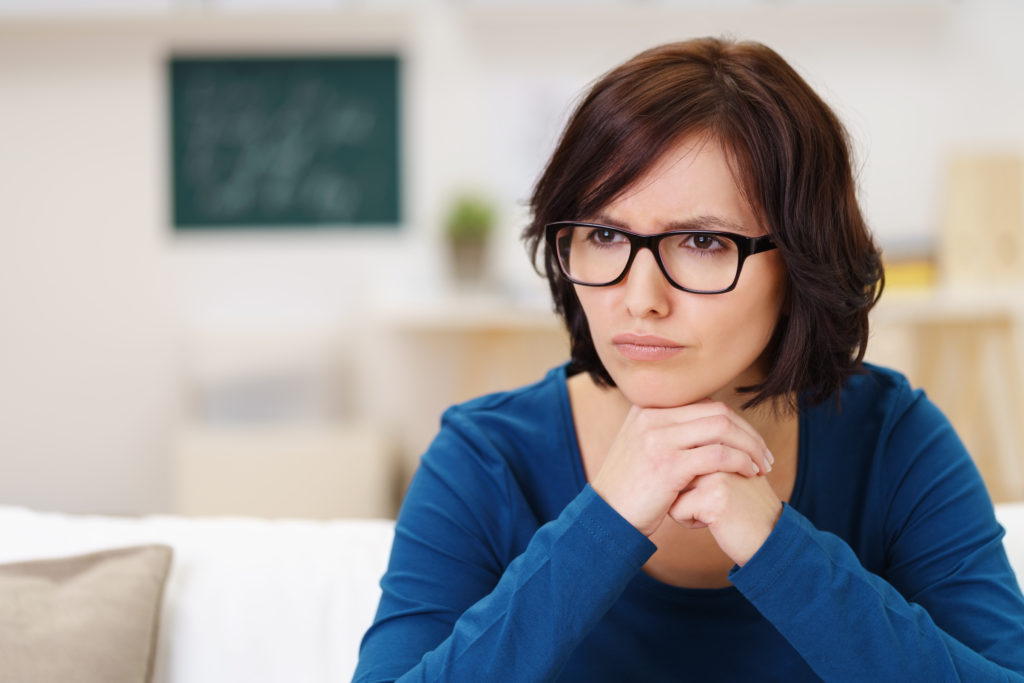 Pensive Woman Wearing Eyeglasses, Sitting on Couch at the Living Room and Looking Away with Chin Resting on her Hands.